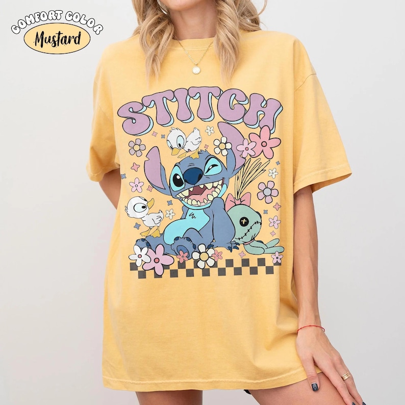 May include: A mustard yellow short-sleeve t-shirt featuring a graphic print of the character Stitch, two small white ducks, and a doll. The text STITCH is printed in purple above the character, surrounded by pink and white flowers and a black checkerboard pattern.