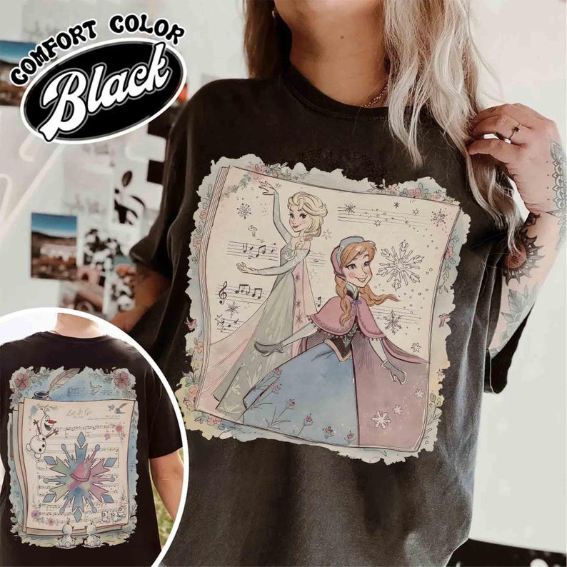 Pode incluir: Camiseta preta Comfort Color com um gr&aacute;fico de duas princesas animadas, notas musicais e detalhes florais. A camisa tamb&eacute;m tem uma imagem menor na parte inferior esquerda das costas com um floco de neve e o texto "Let it go".