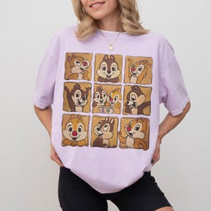 May include: A light purple t-shirt featuring a grid of nine squares, each showcasing a different cartoon chipmunk character. The chipmunks are in various poses and expressions, with brown and tan fur and red noses. The shirt has short sleeves.