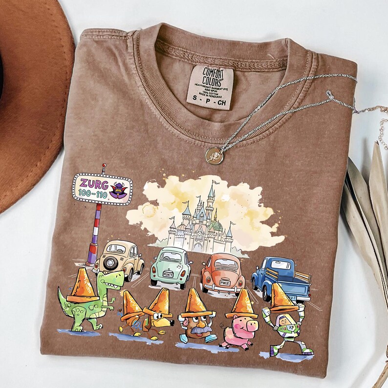 May include: Brown Comfort Colors t-shirt featuring a colorful graphic of cartoon characters in traffic cones, cars, and a castle. The shirt includes the text "Zurg 100-110" on a road sign. A silver necklace with a pendant is also visible.