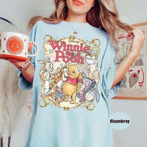 May include: Light blue t-shirt featuring a graphic of Winnie the Pooh and friends. The design includes the text "Winnie the Pooh" in pink and gold lettering, along with illustrations of Pooh, Piglet, Eeyore, and other characters.