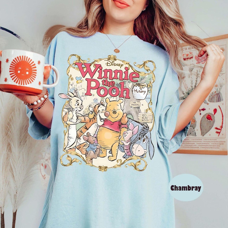May include: Light blue t-shirt featuring a graphic of Winnie the Pooh and friends. The design includes the text "Winnie the Pooh" in pink and gold lettering, along with illustrations of Pooh, Piglet, Eeyore, and other characters.