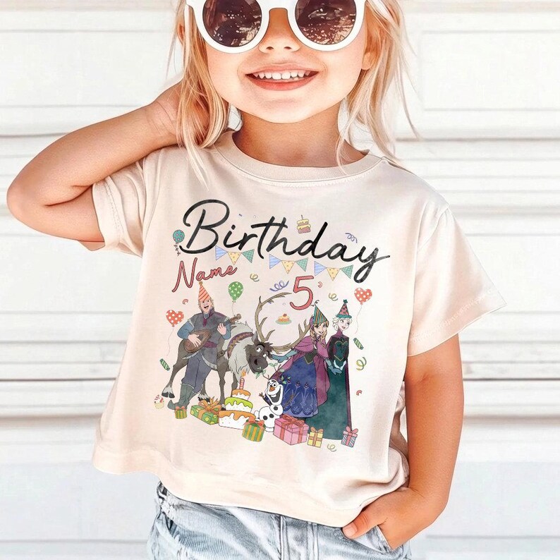 May include: A beige t-shirt with a colorful graphic featuring the word "Birthday" and the number "5". The graphic includes cartoon characters, a reindeer, and birthday cakes. The shirt has short sleeves.