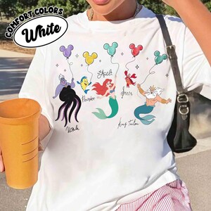 Puede incluir: Camiseta blanca con un gr&aacute;fico colorido que presenta personajes de Disney sosteniendo globos con forma de Mickey Mouse. El dise&ntilde;o incluye a Ariel, Flounder, Sebasti&aacute;n, el Rey Trit&oacute;n y &Uacute;rsula. La camiseta tiene el texto "Comfort Colors" y "White".