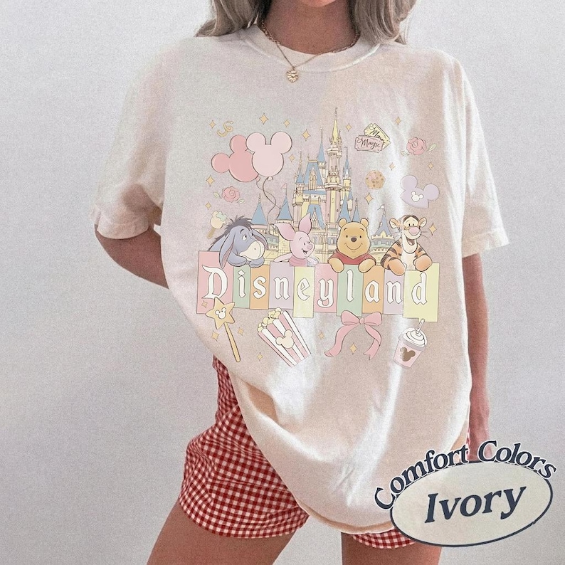 May include: Ivory-coloured t-shirt featuring a Disneyland graphic with Winnie the Pooh characters, a castle, and the word "Disneyland". The shirt has a Comfort Colors label.
