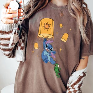 May include: A brown t-shirt featuring a graphic of a blue alien character hanging from a large yellow lantern with a sun design. A small green lizard character is attached to the alien's foot, surrounded by smaller floating yellow lanterns and stars.