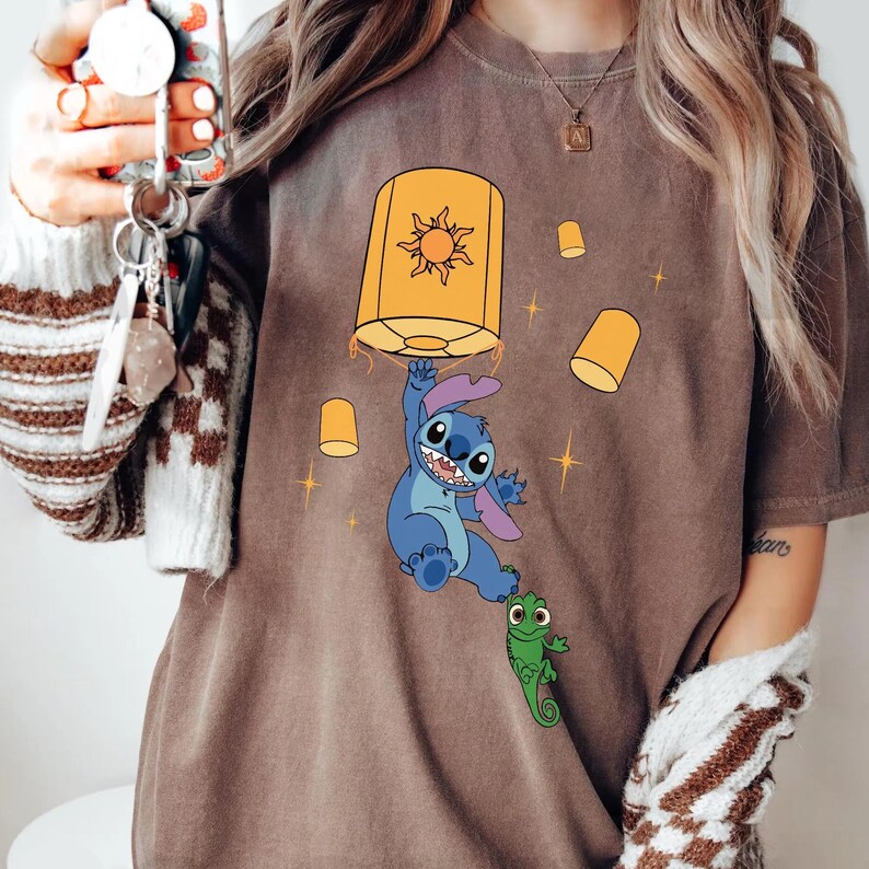 May include: A brown t-shirt featuring a graphic of a blue alien character hanging from a large yellow lantern with a sun design. A small green lizard character is attached to the alien's foot, surrounded by smaller floating yellow lanterns and stars.