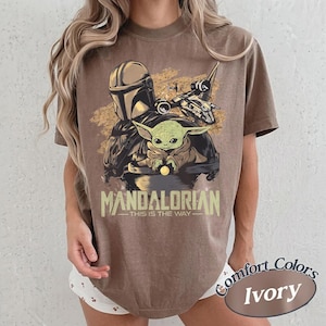 May include: Brown t-shirt featuring a graphic of The Mandalorian, Grogu, and a ship. The text "MANDALORIAN - THIS IS THE WAY" is printed below the image. The shirt is labeled "Comfort Colors Ivory".