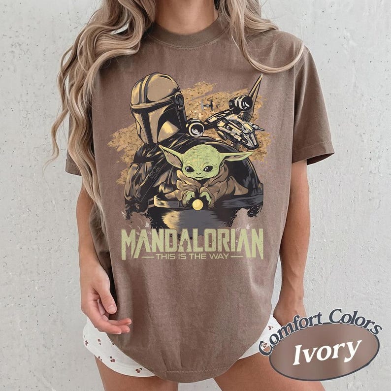 May include: Brown t-shirt featuring a graphic of The Mandalorian, Grogu, and a ship. The text "MANDALORIAN - THIS IS THE WAY" is printed below the image. The shirt is labeled "Comfort Colors Ivory".