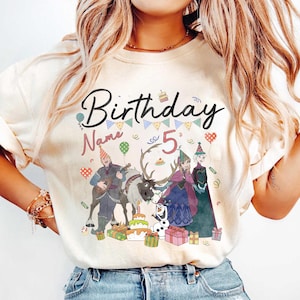 May include: A cream-colored t-shirt featuring a colorful graphic design. The design includes the word "Birthday" in black script, the number "5", and cartoon characters. The design also includes a cake, presents, and balloons.
