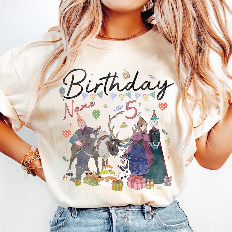 May include: A cream-colored t-shirt featuring a colorful graphic design. The design includes the word "Birthday" in black script, the number "5", and cartoon characters. The design also includes a cake, presents, and balloons.