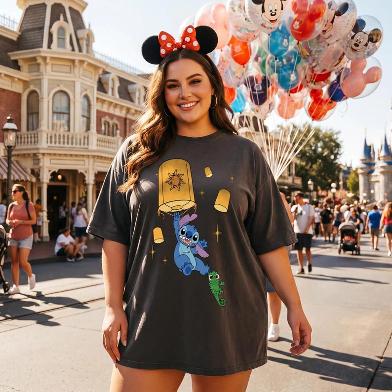 May include: A dark gray t-shirt featuring a graphic print of the blue character Stitch holding a large yellow floating lantern, with a small green chameleon character below and several smaller lanterns and star shapes surrounding them.