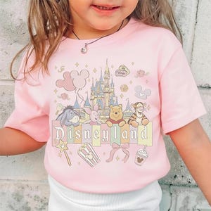 May include: Pale pink t-shirt with a Disneyland design. The graphic includes cartoon characters such as Winnie the Pooh, Tigger, and Eeyore, a castle, balloons, and the word "Disneyland". Additional graphics include a wand, popcorn, a bow, and a drink.