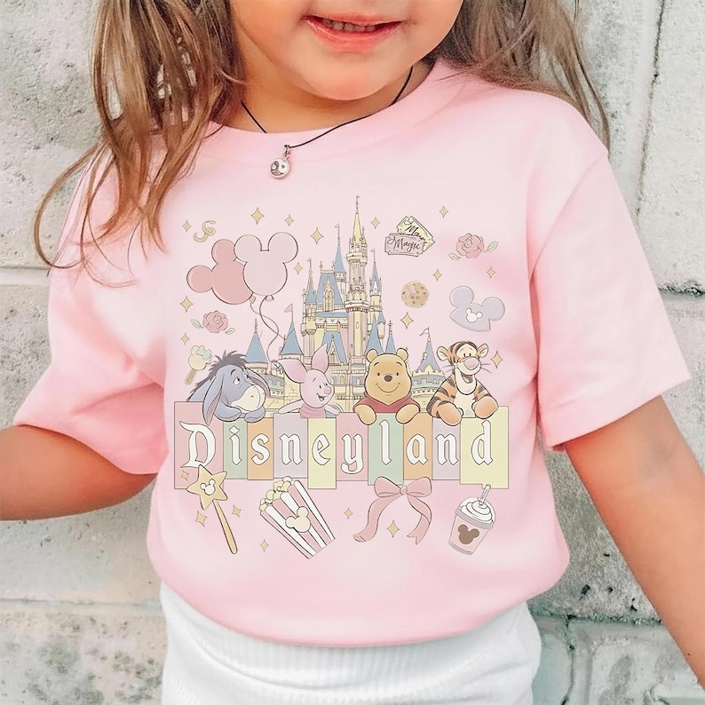 May include: Pale pink t-shirt with a Disneyland design. The graphic includes cartoon characters such as Winnie the Pooh, Tigger, and Eeyore, a castle, balloons, and the word "Disneyland". Additional graphics include a wand, popcorn, a bow, and a drink.