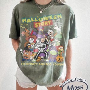 May include: Olive green t-shirt with a Halloween-themed graphic featuring cartoon characters and the text "HALLOWEEN STORY TO INFINITY AND BOO YOUND!". The shirt is a Comfort Colors Moss colour.