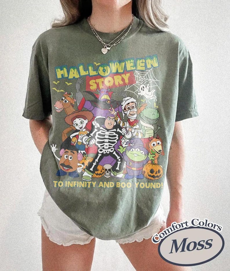 May include: Olive green t-shirt with a Halloween-themed graphic featuring cartoon characters and the text "HALLOWEEN STORY TO INFINITY AND BOO YOUND!". The shirt is a Comfort Colors Moss colour.