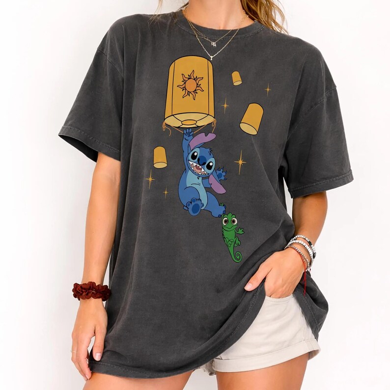 May include: A charcoal gray short-sleeve t-shirt featuring a graphic print of a blue alien character hanging from a yellow lantern with a sun symbol. A small green lizard is positioned below the alien, surrounded by smaller yellow lanterns and stars.