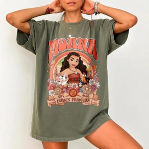 Op de afbeelding: Olijfgroen t-shirt met een afbeelding van Moana en de tekst "Moana" in oranje. Het ontwerp bevat een regenboog, bloemen en de tekst "Est 2016 Disney Princess".