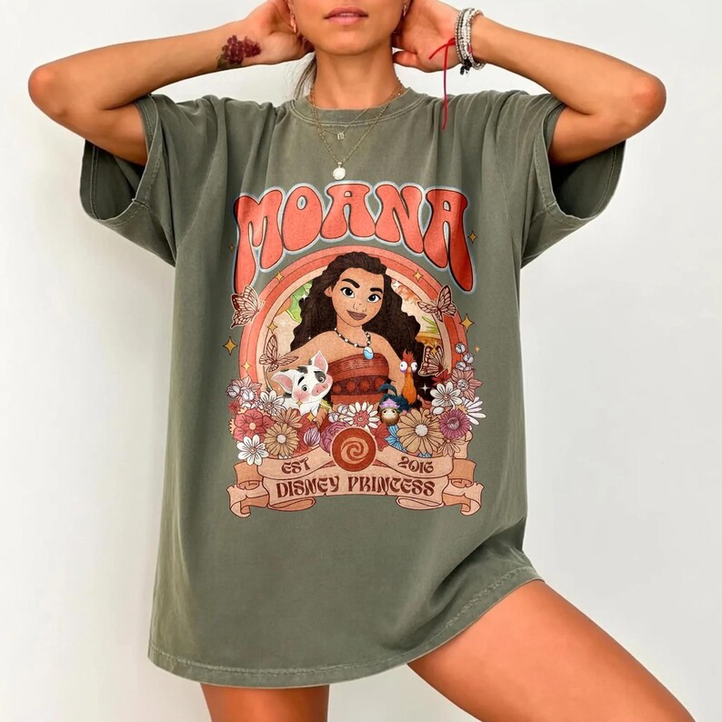 Op de afbeelding: Olijfgroen t-shirt met een afbeelding van Moana en de tekst "Moana" in oranje. Het ontwerp bevat een regenboog, bloemen en de tekst "Est 2016 Disney Princess".