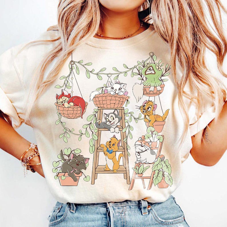May include: Cream-colored sweatshirt featuring a whimsical illustration of various cartoon cats in hanging baskets and pots, surrounded by green vines. The cats are in different poses, some on a wooden ladder, adding a playful touch to the design. The overall theme is a charming cat and plant motif.