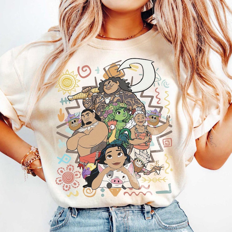 May include: A cream-colored t-shirt featuring a colorful graphic print of animated characters including a large man with tattoos, a woman with long dark hair, an elderly woman, a small green creature, a rooster, and a pig, surrounded by tribal symbols.