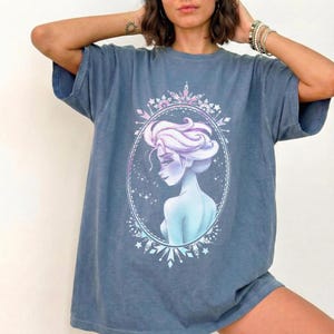 May include: A blue t-shirt featuring a portrait of a woman with light blue skin and purple hair, framed by a decorative oval with snowflake and star accents. The shirt has short sleeves and a relaxed fit.