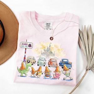 May include: A light pink Comfort Colors t-shirt featuring a whimsical design. The graphic includes cartoon cars, characters, and a castle. A necklace with a pendant and a brown hat are also visible.
