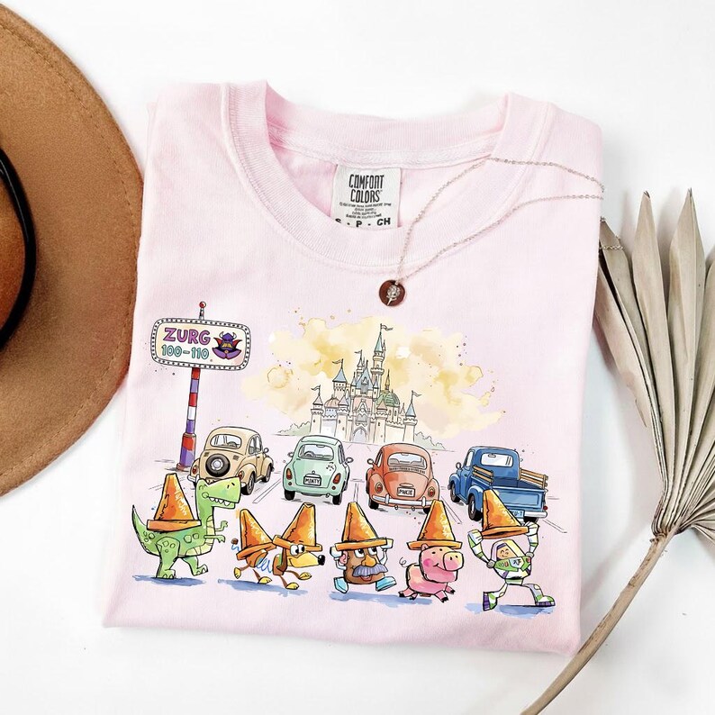 May include: A light pink Comfort Colors t-shirt featuring a whimsical design. The graphic includes cartoon cars, characters, and a castle. A necklace with a pendant and a brown hat are also visible.