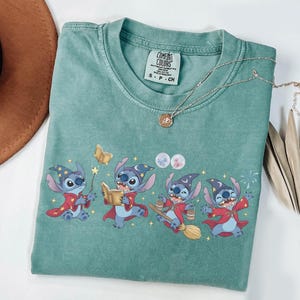 May include: A seafoam green t-shirt featuring a graphic of Stitch from Lilo & Stitch dressed as a wizard. Stitch is wearing a wizard hat and red robe. A gold necklace with a pendant is also visible.