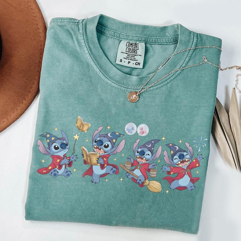 May include: A seafoam green t-shirt featuring a graphic of Stitch from Lilo & Stitch dressed as a wizard. Stitch is wearing a wizard hat and red robe. A gold necklace with a pendant is also visible.