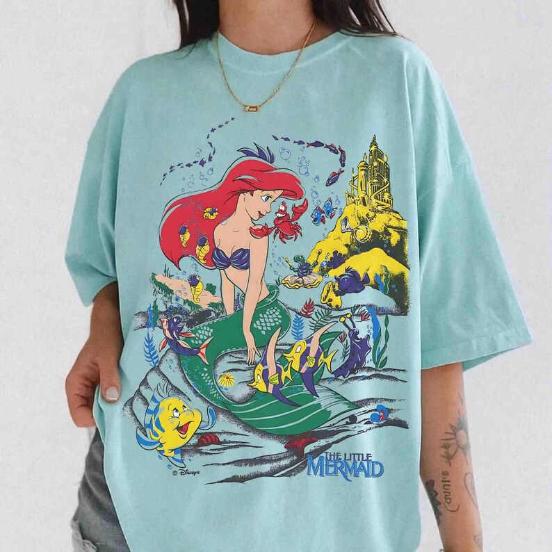 K&ouml;nnte beinhalten: Hellblaues T-Shirt mit einem farbenfrohen Aufdruck von Arielle aus Disneys Arielle, die Meerjungfrau. Das Design zeigt Arielle, Fabius, Sebastian und ein Schloss. Das Shirt hat kurze &Auml;rmel und eine lockere Passform.