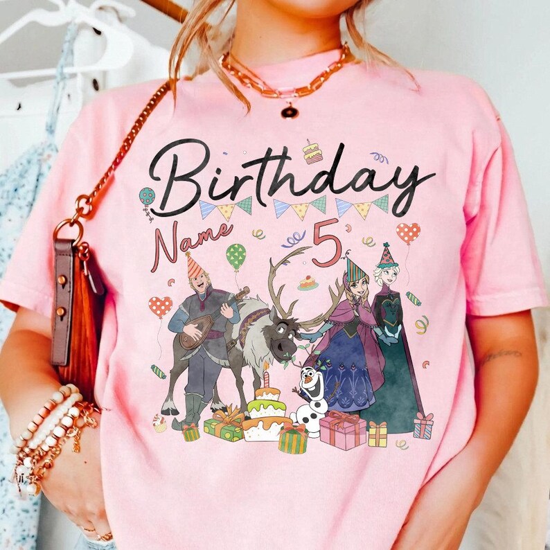 May include: Pink t-shirt with a graphic featuring the word "Birthday" and the number "5". The design includes cartoon characters, a reindeer, a snowman, and birthday gifts. The shirt has a crew neck.