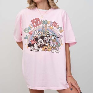 May include: A light pink t-shirt featuring a colorful graphic of Disney characters, including Mickey Mouse, Minnie Mouse, Donald Duck, Goofy, and Pluto. The text "Walt Disney World" is arched above the characters in a rainbow design.
