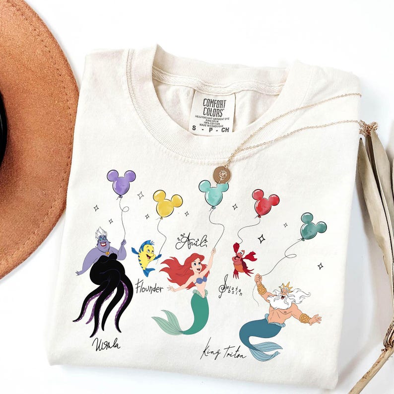 Puede incluir: Camiseta color crema con personajes de La Sirenita. El dise&ntilde;o incluye a &Uacute;rsula, Flounder, Ariel, Sebasti&aacute;n y el Rey Trit&oacute;n, cada uno sosteniendo un globo con forma de Mickey Mouse. La camiseta tiene la etiqueta de la marca Comfort Colors.