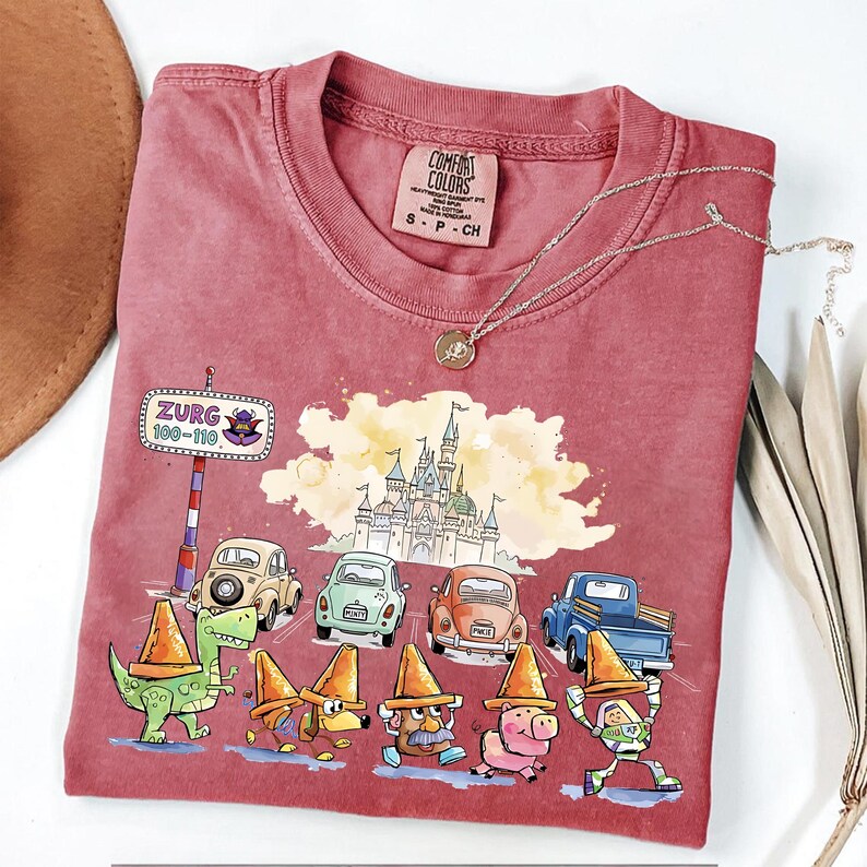 May include: A maroon Comfort Colors t-shirt featuring a graphic of cartoon characters, cars, and a castle. The shirt has the text "Zurg 100-110" on a road sign. A silver necklace with a pendant is also visible.