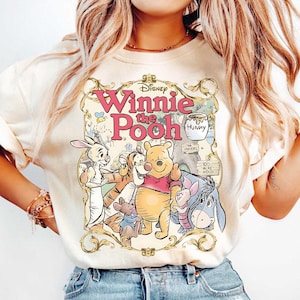 May include: Cream-colored sweatshirt featuring a graphic of Winnie the Pooh and friends. The design includes the text "Winnie the Pooh" in pink, surrounded by a decorative frame. Characters include Pooh, Piglet, Tigger, Eeyore, Rabbit, and Roo.