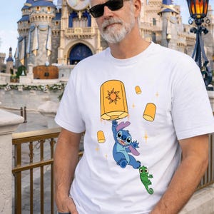 May include: A white short-sleeved t-shirt featuring a graphic print of the blue character Stitch holding onto a large orange floating lantern with a sun design, accompanied by a small green chameleon and several smaller floating lanterns.