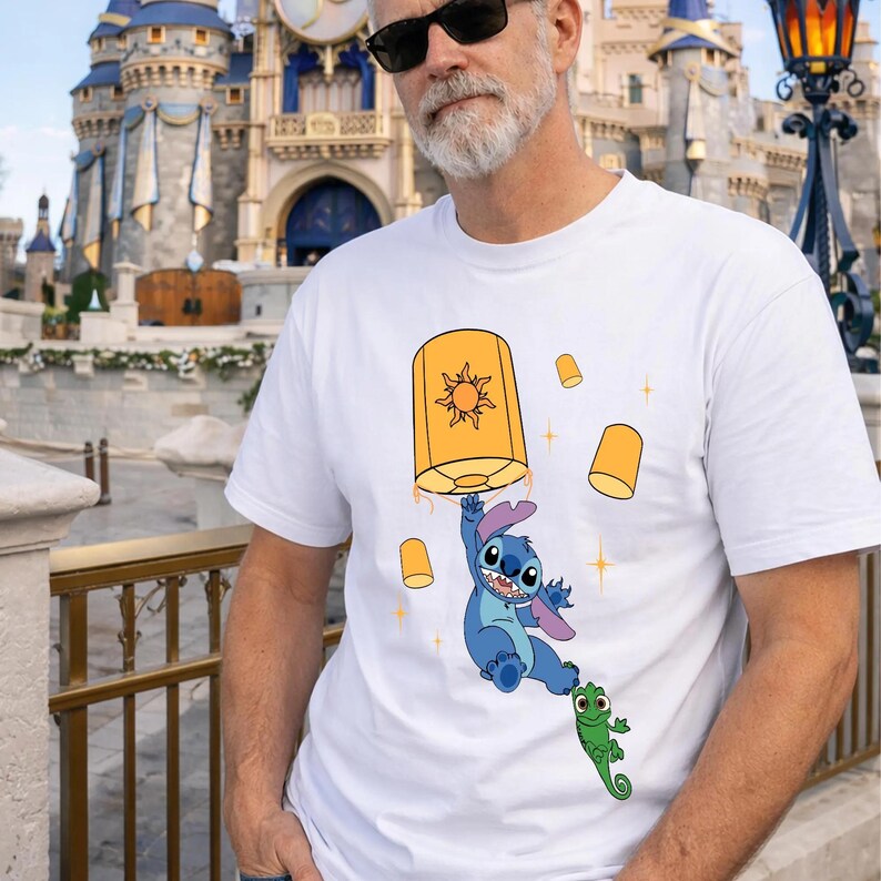 May include: A white short-sleeved t-shirt featuring a graphic print of the blue character Stitch holding onto a large orange floating lantern with a sun design, accompanied by a small green chameleon and several smaller floating lanterns.