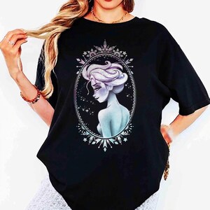 May include: Black t-shirt featuring a detailed illustration of a woman with light blue skin and white hair, framed within an oval border. The design has a celestial theme with stars and snowflake accents, creating a fantasy aesthetic.