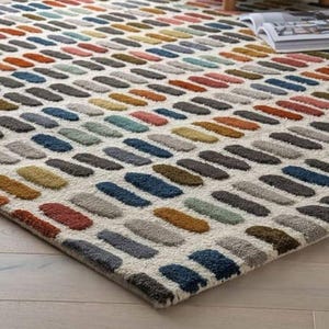 Könnte beinhalten: Ein rechteckiger Teppich mit einem sich wiederholenden Muster aus farbigen, länglichen Formen in Rot-, Orange-, Gelb-, Grün-, Blau- und Grautönen. Der Teppich hat einen cremefarbenen Hintergrund und liegt auf einem hellen Holzboden. Ein gelbes Stuhlbein ist sichtbar.