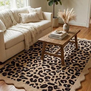 Könnte beinhalten: Ein Wohnzimmer mit beigem Sofa, Kissen und einer Fransen-Decke. Ein Holztisch zeigt Bücher und eine Vase mit getrockneten Pflanzen. Ein Teppich mit Leopardenmuster und gewelltem Rand liegt auf dem Holzboden.