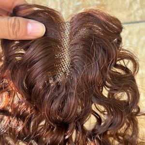 Op de afbeelding: Een close-up van een bruin haarstuk met golvende, krullende lokken. Het haarstuk heeft een mesh basis met kleine, goudkleurige bevestigingen. Het haar is een rijke, donkerbruine kleur met roodachtige highlights en de textuur lijkt zacht en natuurlijk.