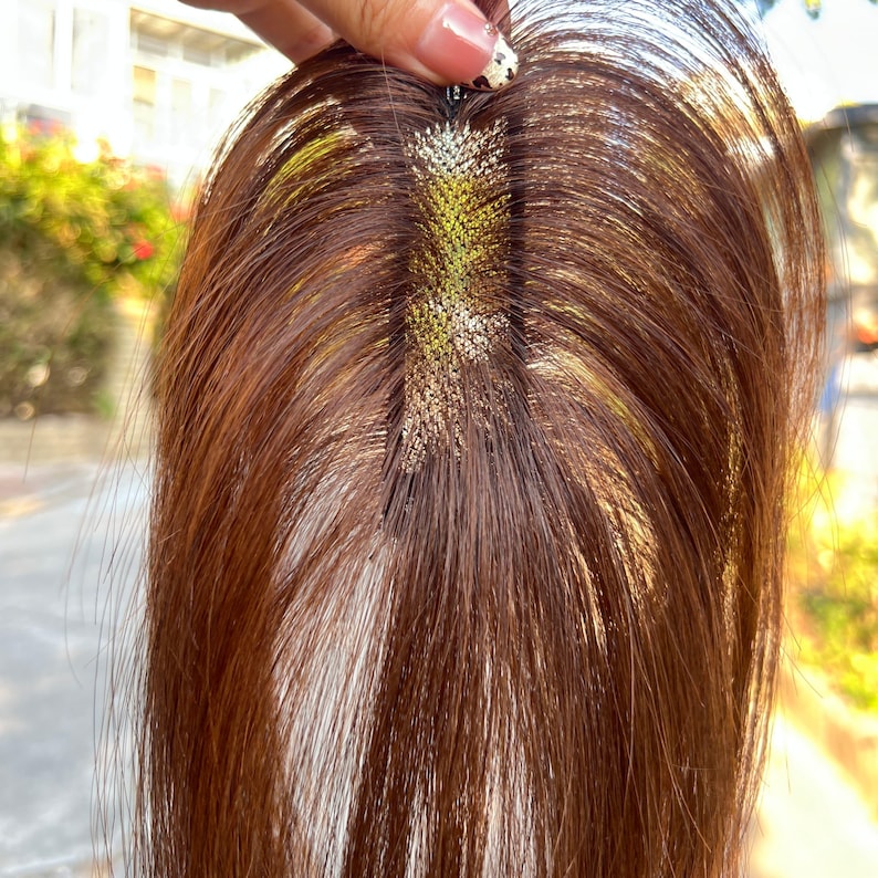May include: A brown hair topper with a clip attachment. The hairpiece has a natural-looking texture and color, with a visible part. The base of the topper features a mesh-like design with a gold-colored accent.
