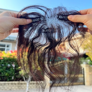 May include: A dark brown hairpiece with a wavy texture, held up. The hairpiece has a mesh base with clips for attachment. The hair appears natural and is styled with loose waves. The hairpiece is designed to add volume.