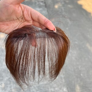 Op de afbeelding: Een bruin haarstuk met een mesh basis en lange, rechte pony. Het haarstuk wordt door een hand omhoog gehouden, waarbij de kleur en textuur van het haar worden getoond. Het haarstuk is ontworpen om volume en stijl aan het haar toe te voegen.