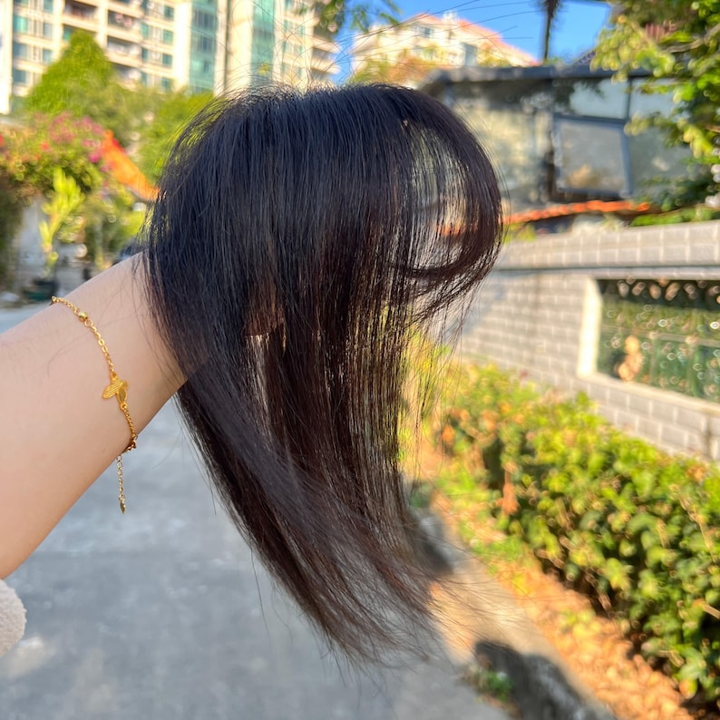 May include: A dark brown hairpiece with long, layered strands. The hairpiece is held up against a blurred outdoor background. A gold bracelet with a butterfly charm is visible on the wrist.