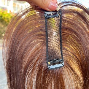 May include: A hair topper with reddish-brown hair and a black mesh base. The topper has small, metal clips for secure attachment. The hair appears to be layered, with highlights adding dimension. The hairpiece is designed to add volume and coverage.