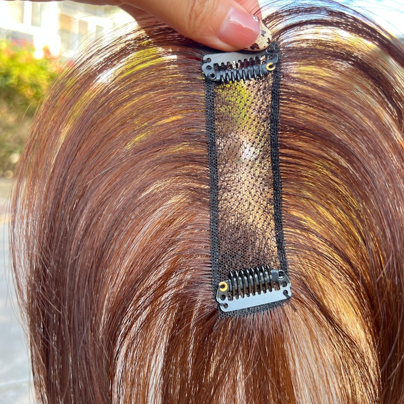 May include: A hair topper with reddish-brown hair and a black mesh base. The topper has small, metal clips for secure attachment. The hair appears to be layered, with highlights adding dimension. The hairpiece is designed to add volume and coverage.