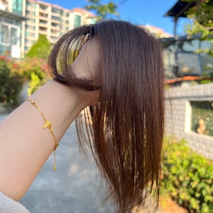 May include: A gold-colored bracelet with a small bird charm is worn on a wrist. The bracelet is delicate, with a thin chain and small beads. The background is blurred, with greenery and buildings visible.