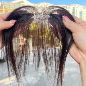 May include: A dark brown hairpiece with a fine mesh base, held up to the light. The hairpiece appears to be a fringe or bangs extension, designed to blend with natural hair. The hair strands are long and straight, with a slight sheen.